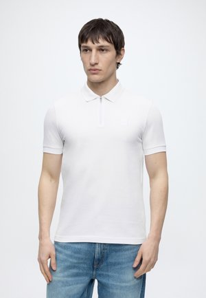 Ung mand iført en hvid kortærmet polo med lynlåslukning ved kraven og blå jeans, stående foran en ensfarvet lys baggrund.