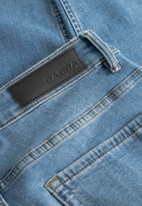 Lichtblauwe denim met een gestructureerde afwerking, voorzien van een zwarte leren patch met het label "GABBA" en contrasterende oranje stiksels.