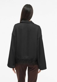 Svart bluse i glatt stoff, med blonder i kanten nederst, vide ermer og klassisk krage.