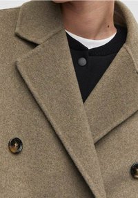 Cappotto di lana doppiopetto in verde oliva. Presenta revers marcati, due grandi bottoni neri e una finitura in tessuto testurizzato. Indossato sopra un maglione nero.