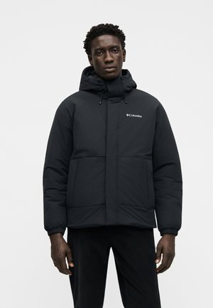 Mann trägt eine schwarze Columbia Steppjacke mit Kapuze und Fronttaschen und steht vor einem schlichten hellgrauen Hintergrund.