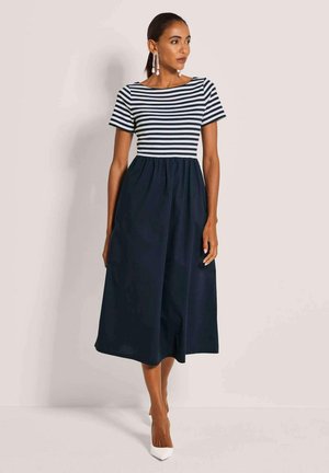 Frau trägt ein kurzärmliges Kleid mit schwarzen und weißen Streifen, kombiniert mit einem schwarzen Midirock und weißen spitzen High Heels, steht still.