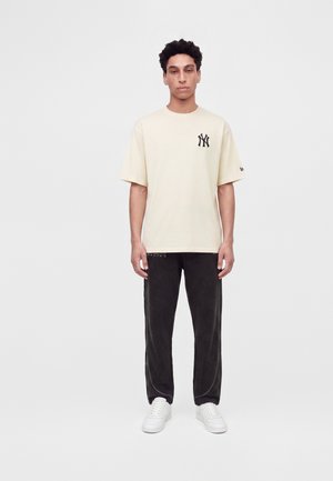 Мъж, стоящ, облечен с бежова тениска на New York Yankees, черни панталони с текст "RACING" и бели маратонки на светъл едноцветен фон.