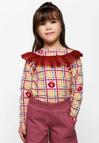 Langarmtop met een multikleurig geruit patroon met appelsymbolen en een rode gerimpelde kraag, gecombineerd met kastanjebruine high-waisted broeken.