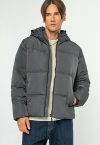 Přehnaná šedá bunda s kapucí, která m�á vodorovné švy, zapínání na zip a dvě boční kapsy, nošená přes světlý top a džíny.