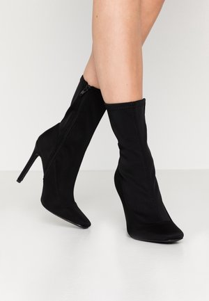 Schwarze kniehohe Stiefeletten mit hohen Absätzen und seitlichen Reißverschlüssen, getragen an übereinandergeschlagenen Beinen vor hellem, einfarbigem Hintergrund.