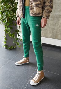 Green Nike sweatpants with an elastic waistband, paired with tan high-top Converse shoes featuring white soles, set against a modern background.
