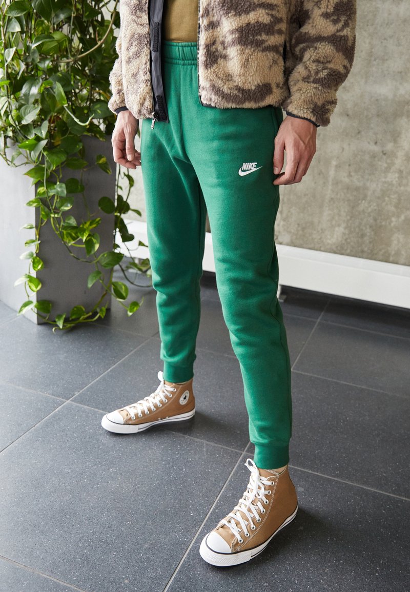 Green Nike sweatpants with an elastic waistband, paired with tan high-top Converse shoes featuring white soles, set against a modern background.
