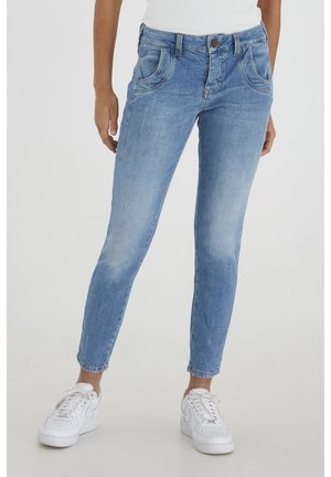 Personne portant un jean skinny bleu clair et des baskets blanches, debout devant un fond clair uni.