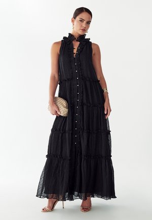 Woman wearing a sleeveless black ruffled maxi dress with gold buttons, holding a woven gold clutch, and wearing gold high-heeled sandals.
