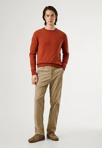 Rozsda színű pulóver, bézs nadrág és barna cipő. A pulóver kerek nyakkal rendelkezik; a nadrág egyenes szabású zsebekkel.