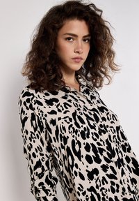 Schwarz-beige Bluse mit Leopardenmuster, durchgehender Knopfleiste und langen Ärmeln mit aufgerollten Manschetten. Sanfte Stoffstruktur.