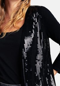 Cardigan nero con paillettes, dalla superficie testurizzata, caratterizzato da una vestibilità ampia e maniche lunghe, indossato sopra un top nero.