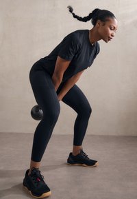 Schwarz athletic Shirt, dunkle Leggings mit Nahtdetails und schwarze Trainingsschuhe mit Gummisohlen. Der Griff der Kettlebell ist hervorgehoben.