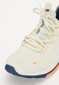 Light cream running shoe with wave pattern texture, cushioned sole, blue outsole, elastic laces, and orange accent on heel.
