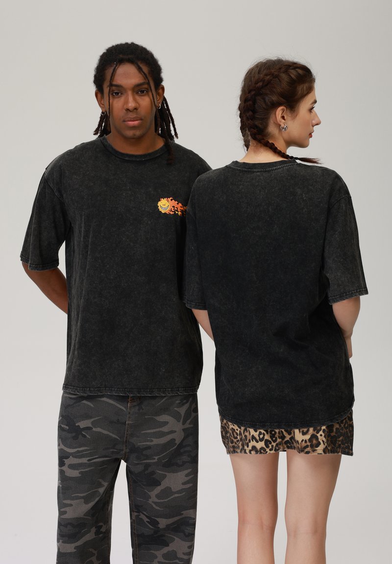 T-shirt gráfica oversized, de algodão e preta, com um design colorido de rosto sorridente na parte da frente. Tecido texturizado e desbotado, com mangas arregaçadas.