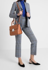 Costume à carreaux gris avec un blazer ajusté et un pantalon court, associé à un col roulé bleu et des ballerines à talons ouverts noires. Sac à main marron avec des accents en métal.