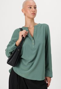 Top verde a maniche lunghe con scollatura henley, tre bottoni dorati e maniche arrotolabili. Abbinato a una borsa nera. Texture liscia.