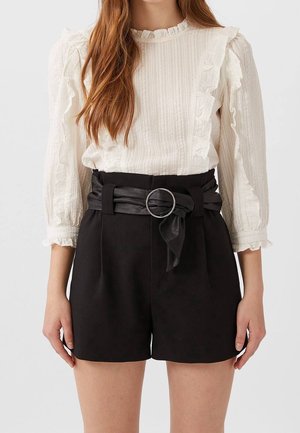 Witte textuur blouse met gerimpelde mouwen en een zwarte high-waisted short met een gestrikt stoffen riem en een zilveren ringaccent.