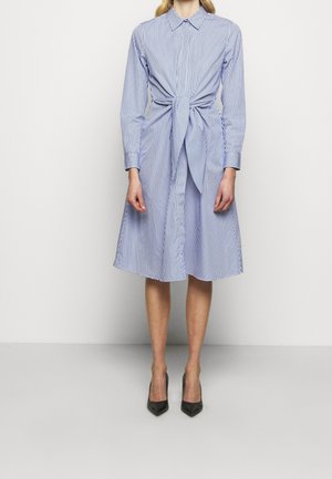 Femme portant une robe à manches longues rayée verticalement bleu et blanc avec un nœud devant et des talons noirs à bout pointu.