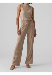 Beżowy kombinezon w plisowanym stylu z bezrękawowym krojem, wysokim kołnierzem, elastycznym pasem i szerokimi nogawkami. Noszony z płaskimi sandałami na paski.