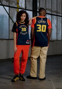 Due persone indossano maglie sportive. La donna ha una maglia nera con "GOLDEN STATE" e "30" in giallo, abbinata a pantaloni rossi. L'uomo indossa una maglia blu simile, con "CURRY" e "30" in giallo, insieme a pantaloni color cammello.