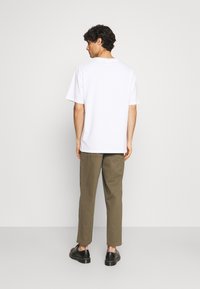Camiseta de algodón blanca de manga corta, combinada con pantalones verdes oliva. El modelo lleva zapatos negros; fondo neutro.