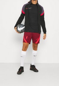 Henkilö seisoo pitäen mustavalkoista jalkapalloa, pukeutuneena Nike-takkiin, Liverpool-shorteihin ja sukkiin, jalassaan urheilukengät.