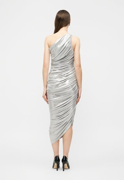 Γυναίκα με ασημένιο φόρεμα μίντι με έναν ώμο και ραβδώσεις, και μαύρες ψηλοτάκουνες γόβες, στέκεται με την πλάτη προς τα εμπρός σε απλό φόντο.