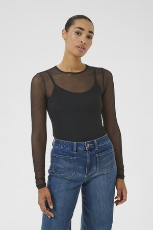 Femme portant un haut noir transparent à manches longues par-dessus un caraco noir, associé à un jean bleu taille haute sur un fond uni.