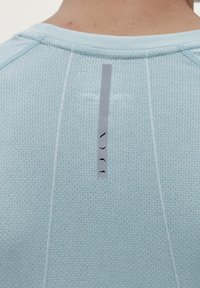 Světle modré sportovní tričko s texturovanou tkaninou. Na zadní straně má vertikální šedou etiketu s minimalistickými černými akcenty.