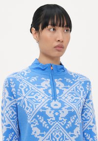 Woman with black hair in a bun wearing a blue and white patterned zip-up sweater, looking slightly to the right against a plain background.