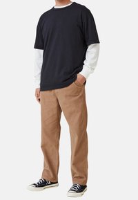 Man som bär en lager på lager svart kortärmad skjorta över en vit långärmad skjorta, bruna kordflortbyxor och svarta sneakers, stående med en hand i fickan.