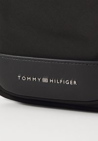 Černá nylonová taška s hladkým koženým dnem, s embosovaným stříbrným logem "TOMMY HILFIGER" a elegantním, minimalistickým designem.