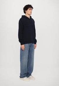 Felpa blu navy con tasca anteriore e logo testurizzato, abbinata a jeans larghi azzurri e sneaker bianche. Vestibilità semplice e rilassata.