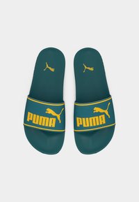 Τσαλακωτές σαμπό σαγιονάρες σε τιρκουάζ χρώμα με κίτρινο λογότυπο Puma και γράμματα στο λουράκι και στην εσωτερική σόλα, τοποθετημένες δίπλα-δίπλα σε λευκό φόντο.