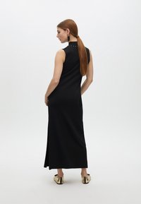 Vestido maxi negro sin mangas con cuello alto y sutiles tachuelas en el collar. Textura suave, ligera abertura en el costado, combinado con calzado dorado.