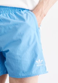 Svetle modré sportovní šortky s elastickým pasem, bílé logo v dolním levém rohu a bílé boční pruhy. Texturovaná tkanina s hladkým povrchem.