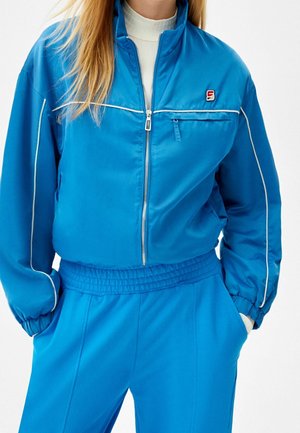 Femme portant une veste zippée bleu vif avec un liseré blanc et un pantalon assorti à taille élastique, mains dans les poches, col roulé blanc en-dessous.