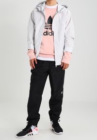 Ljusgrå zip-up jacka med huva, rosa sweatshirt med svart logotyp, svarta byxor med vita ränder och svarta sportskor med detaljer.