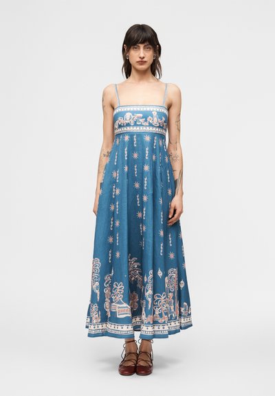 Donna con capelli scuri che indossa un vestito blu alla caviglia con spalline sottili e motivi intricati bianchi e rosa, abbinato a scarpe rosse con lacci.