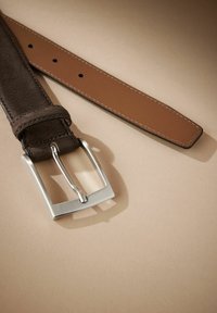 Brun læderbælte med en ruskindsstruktur, der har en rektangulær metalspænde i sølv. Stropen har flere justeringshuller.