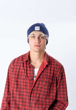 Junger Mann mit blondem Haar, der eine blaue Beanie und ein rotes kariertes Hemd trägt, schaut mit neutralem Gesichtsausdruck auf weißem Hintergrund geradeaus.