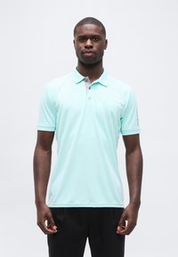 Νέος άνδρας που στέκεται όρθιος φορώντας ανοιχτό μπλε πουλόβερ πόλο και μαύρο παντελόνι μπροστά σε απλό λευκό φόντο.