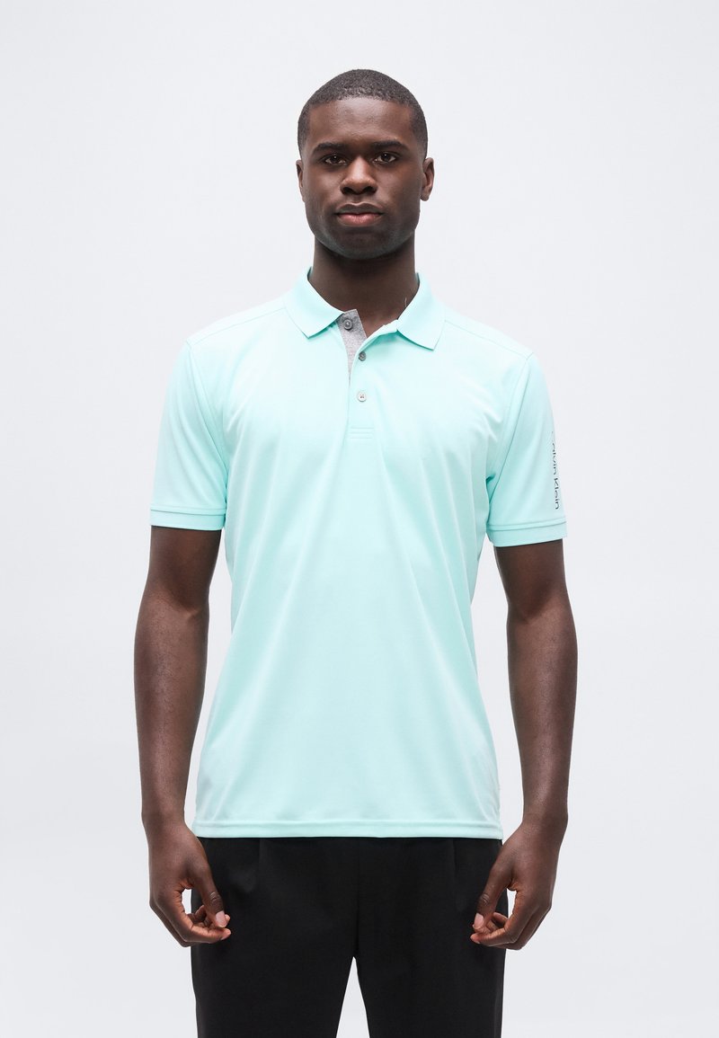 Νέος άνδρας που στέκεται όρθιος φορώντας ανοιχτό μπλε πουλόβερ πόλο και μαύρο παντελόνι μπροστά σε απλό λευκό φόντο.