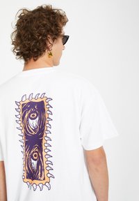 Hvid bomulds T-shirt med et stort grafisk motiv på ryggen i lilla og orange farver, der viser øjne og takkede kanter.