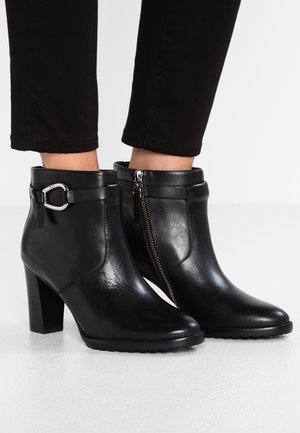 Bottines en cuir noir avec talons blocs, fermeture éclair latérale et sangle à boucle portées avec un pantalon noir sur une surface blanche.