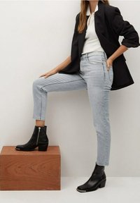 Botas negras hasta el tobillo con un acabado brillante, combinadas con jeans rectos de color azul claro y un blazer negro sobre una camiseta blanca, posadas sobre un bloque de madera.