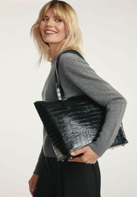 Czarna torba na ramię z wytłoczonym wzorem krokodyla, z dwoma uchwytami, w usztywnionym kształcie, z płaskim dnem i metalowymi nóżkami. Modelka nosi szary sweter.