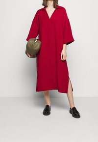 Femme portant une robe midi rouge ample avec une fente latérale, tenant un sac vert olive, et portant des mocassins noirs brillants.